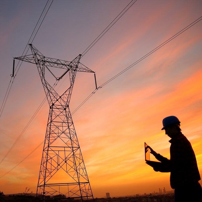 Ein Strommast steht vor einem farbenfrohen Sonnenuntergang. Im Vordergrund ist die Silhouette einer Person mit einem Laptop, die in Richtung des Mastes schaut. Die Szene strahlt eine Atmosphäre der Technologie und Energie aus.