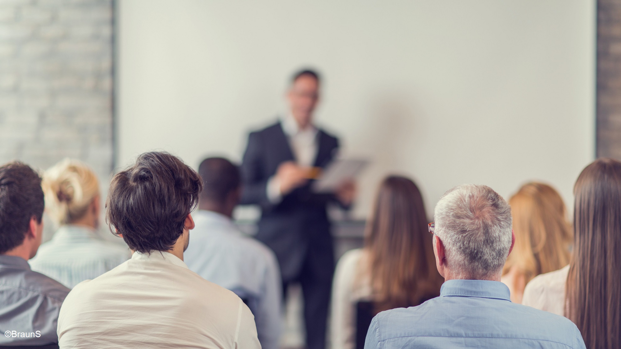 A diverse group of people is seated, facing a speaker in a business suit. The speaker is positioned in front of a blank presentation screen, holding papers. The audience appears engaged, attentively listening to the presentation.