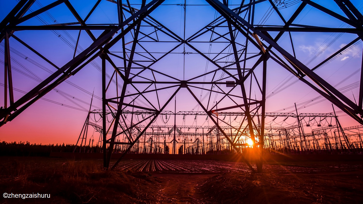 Ein Netz von Hochspannungsmasten steht im Vordergrund, silhouettiert vor einem farbenfrohen Sonnenuntergang. Der Himmel zeigt sanfte Farbtöne von Blau und Lila, während die Sonne am Horizont untergeht. Im Hintergrund sind weitere Masten und elektrische Leitungen sichtbar.