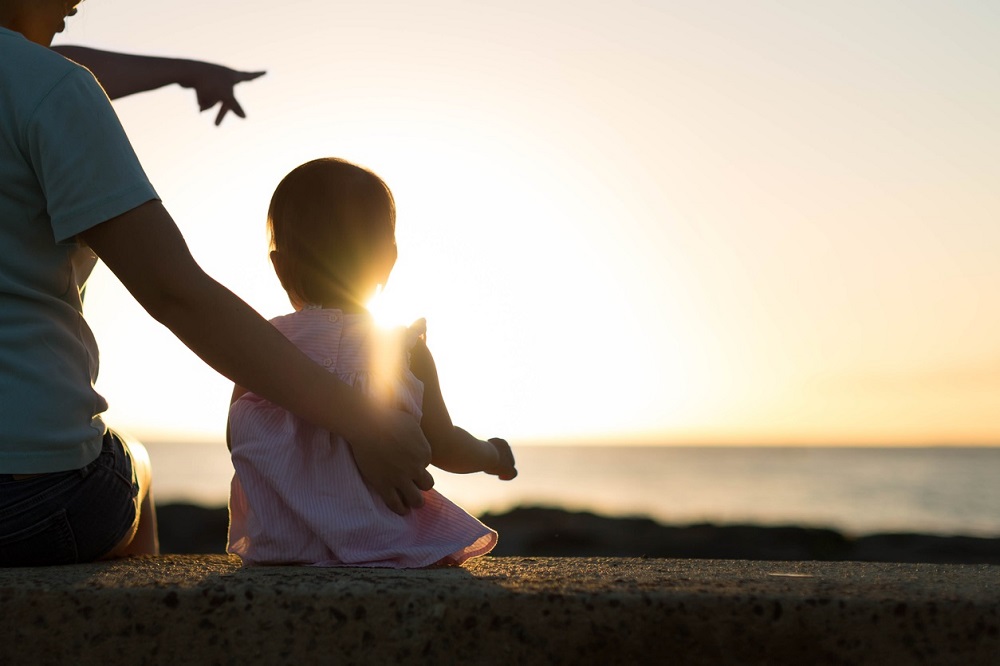 A caregiver sits with a small child, both facing a sunset over the water. The caregiver points towards the horizon, sharing a moment of connection and wonder as the warm sunlight illuminates their silhouettes.