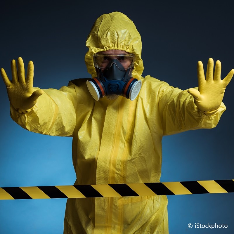 Eine Person in einem gelben Schutzanzug und Atemschutzmaske hält die Hände in einer abwehrenden Geste vor sich. Sie steht hinter einer absperrenden Gelb-Schwarz-Band, was auf eine gefährliche oder kontaminierte Zone hinweist.