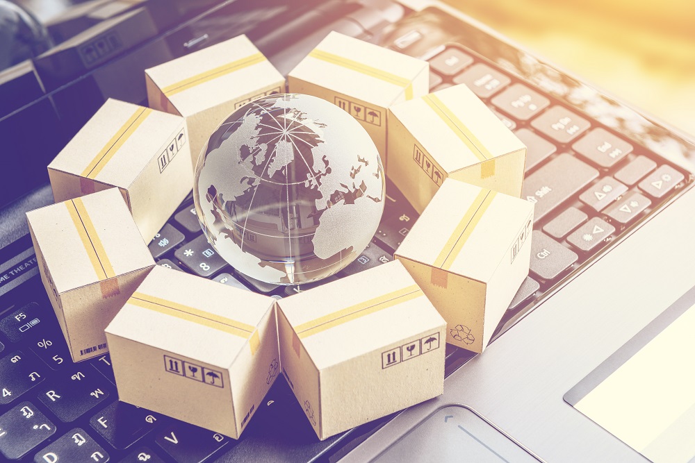 Eine Glaskugel mit einer Weltkarte steht auf einer Laptop-Tastatur, umgeben von mehreren Paketen mit gelben Streifen. Das Bild symbolisiert globalen Versand und E-Commerce.