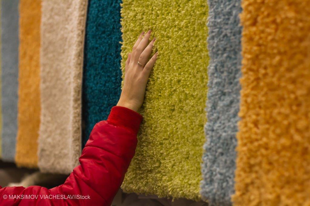 A hand is reaching out to touch a display of colorful, fluffy rugs hanging vertically. The rugs feature a range of soft textures and colors, including green, blue, orange, and yellow. The setting suggests a store or showroom for home textiles.