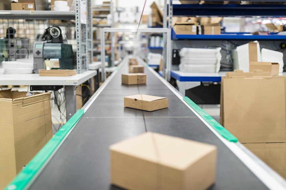 Ein Förderband transportiert mehrere Kartons in einem Lagerraum. Im Hintergrund sind Regale mit weiteren Kartons und Verpackungsmaterialien sichtbar. Das Bild vermittelt den Eindruck eines aktiven Logistikprozesses.