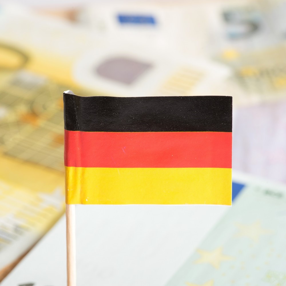 Eine kleine deutsche Flagge in Schwarz-Rot-Gold, die aufrecht auf einem Stab steht. Im Hintergrund sind unscharfe Banknoten sichtbar, die nicht näher beschrieben werden.