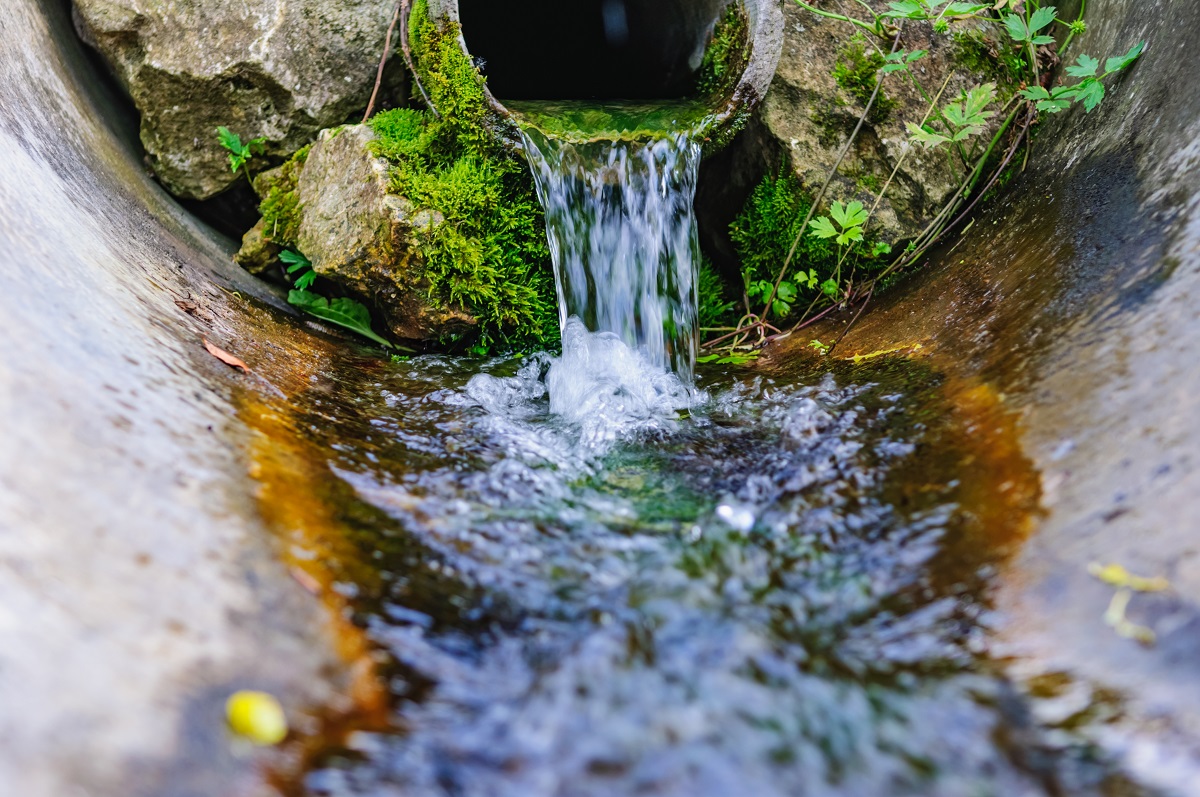 Ein Wasserstrahl fließt aus einem Rohr, umgeben von grünem Moos und Steinen. Das Wasser bildet kleine Wellen und sprudelt ca. in einem betonierten Bereich, der von natürlicher Vegetation umgeben ist. Die Szene vermittelt ein Gefühl von Frische und Natur.
