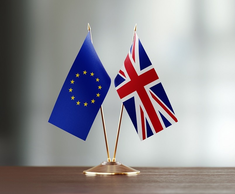 Zwei Flaggen stehen auf einem Tisch: links die europäische Flagge mit einem blauen Hintergrund und gelben Sternen, rechts die britische Flagge, bekannt als Union Jack. Beide Flaggen sind auf kleinen Ständern platziert.