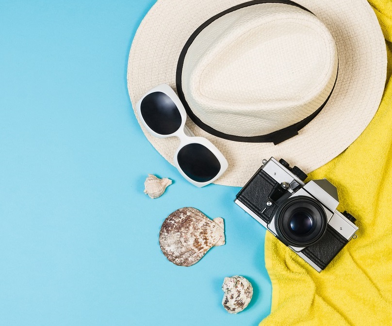 Auf einem blauen Hintergrund liegen ein Strohhut, schwarze Sonnenbrillen, eine Kamera und einige Muscheln. Ein gelbes Handtuch ist ebenfalls zu sehen, das die Szene ergänzt und ein Urlaubsgefühl vermittelt.