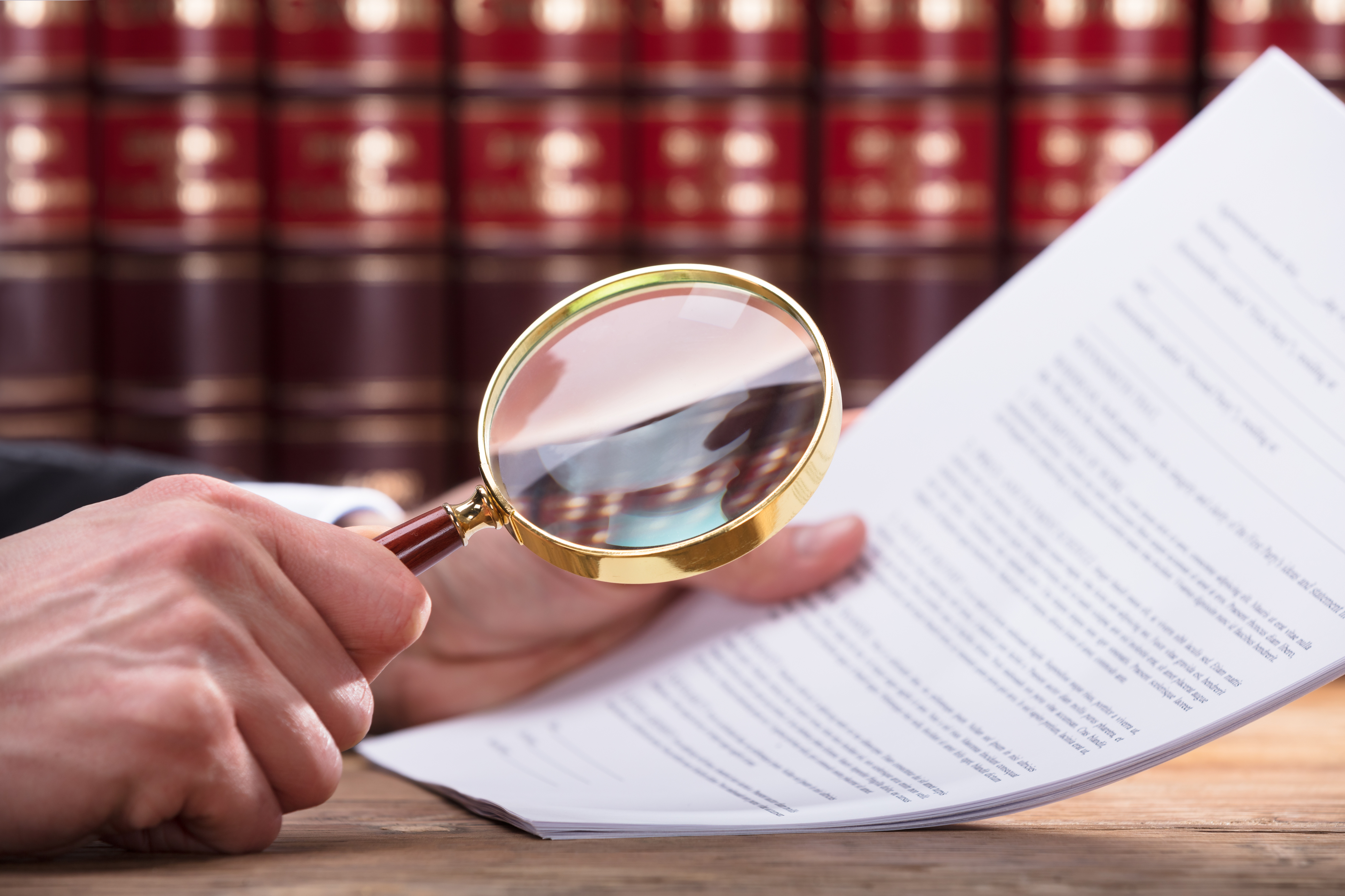 Eine Hand hält eine Lupe über ein Blatt Papier mit Text, während im Hintergrund Bücherregale sichtbar sind. Die Lupe wird verwendet, um Details im Dokument genauer zu betrachten.
