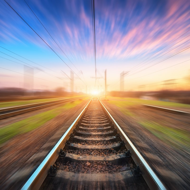 Ein Gleis mit Schienen verläuft in die Ferne, unter einem farbenfrohen Himmel während des Sonnenaufgangs. Die Atmosphäre vermittelt ein Gefühl von Bewegung und Geschwindigkeit, während das Licht am Horizont aufleuchtet.