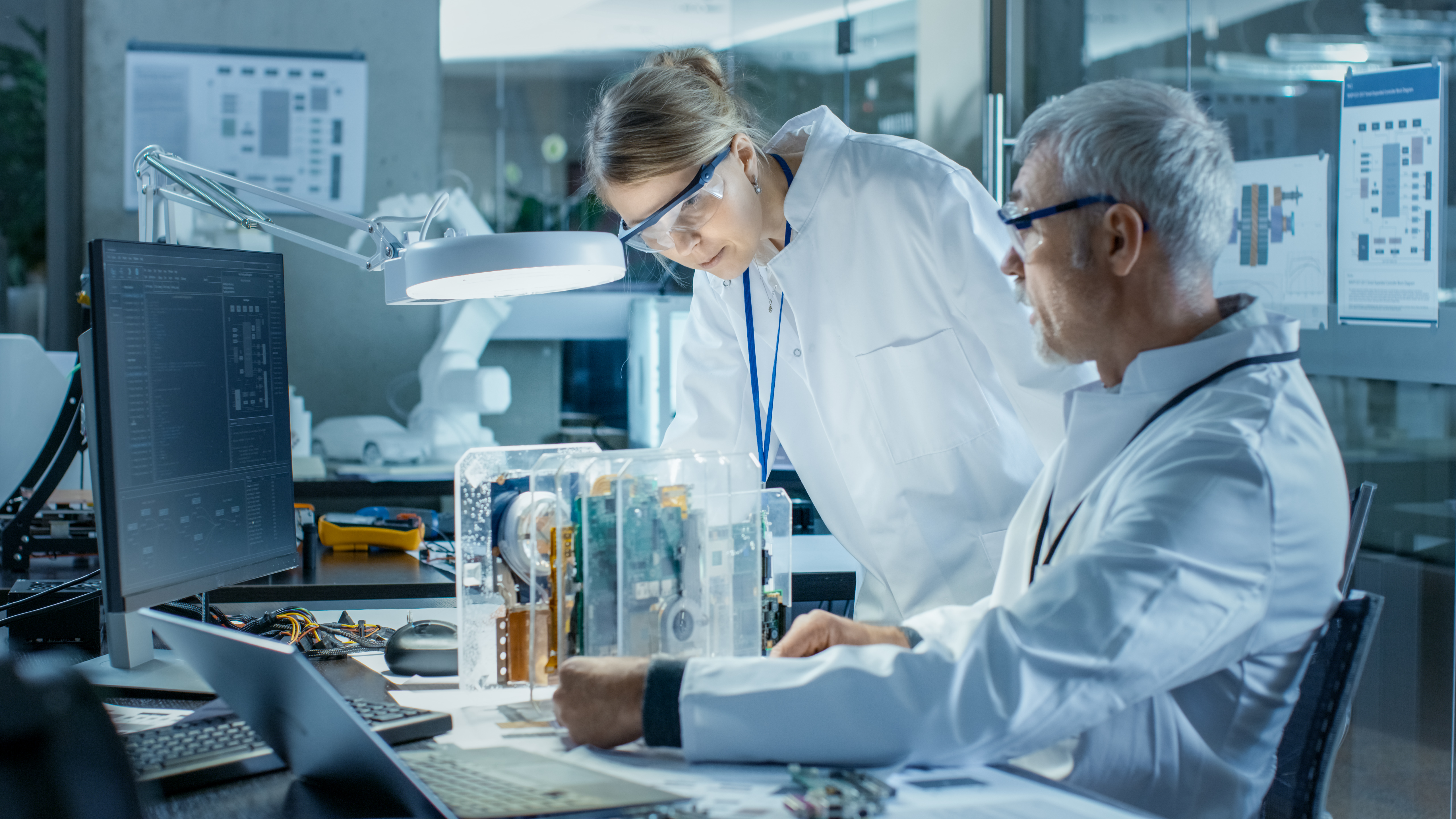 Two scientists in lab coats work together in a modern laboratory. One, focused on a transparent device with electronic components, examines it closely while the other reviews information on a computer monitor. The lab features advanced technology and a workspace filled with various tools and equipment.