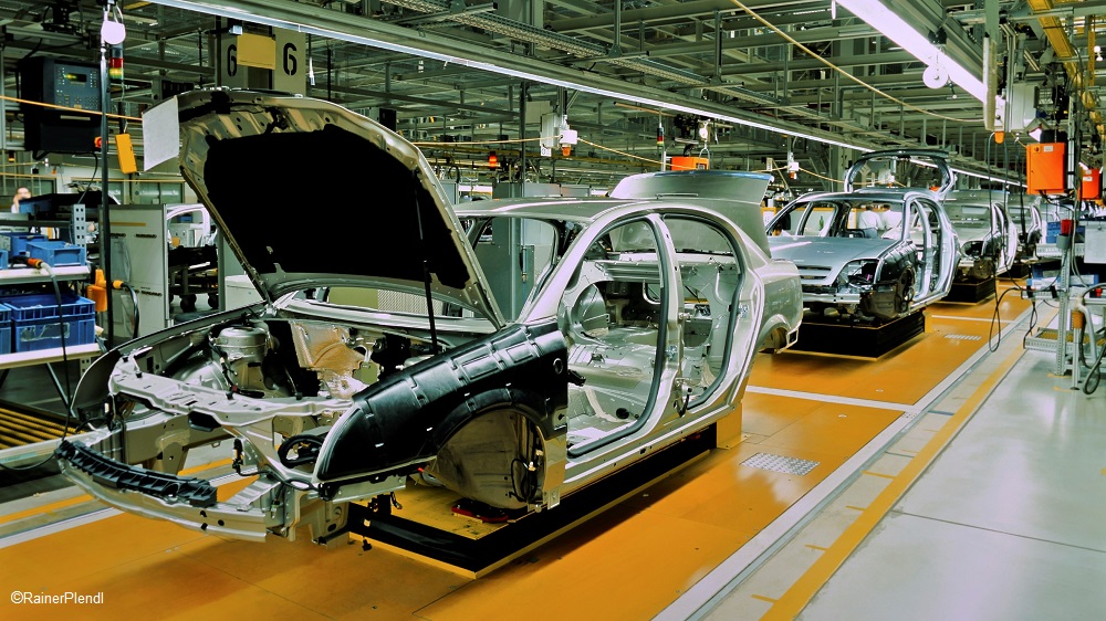 Ein Automobilwerk zeigt mehrere unvollständige Autofahrgestelle auf einem Montageband. Die Wagen sind teils lackiert und ohne Motorhaube. Im Hintergrund sind weitere Fahrgestelle zu sehen, was den industriellen Fertigungsprozess verdeutlicht.