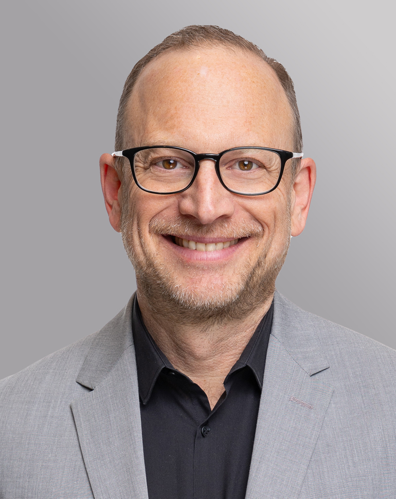 Ein Mann mit Brille lächelt in die Kamera. Er trägt einen grauen Anzug und ein schwarzes Hemd. Der Hintergrund ist grau und hell, wodurch sein Gesicht gut zur Geltung kommt.