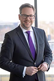 A professional man stands smiling, dressed in a tailored suit with a purple tie. He is positioned indoors with natural light coming through a large window, suggesting a modern office environment. His hands are clasped in front of him, conveying confidence and approachability.