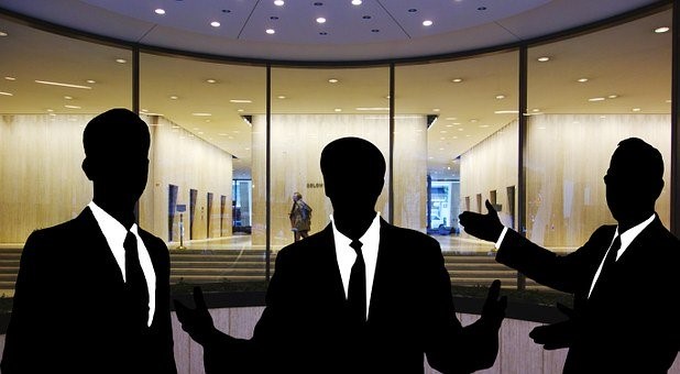 Drei silhouettenhafte Figuren in Anzügen stehen vor einer großen Glastür. Sie scheinen miteinander zu sprechen und gestikulieren. Im Hintergrund ist eine Person, die die Glasfront passiert, sichtbar. Das Bild vermittelt eine geschäftliche Atmosphäre.