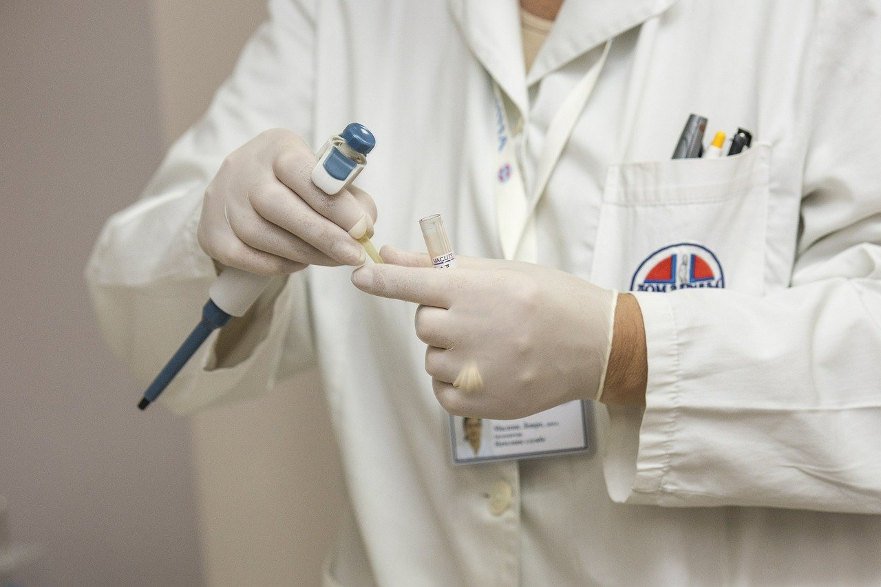 Ein Arzt in einem weißen Kittel hält eine Pipette in der einen Hand und ein Probenröhrchen in der anderen Hand. Beide Hände sind mit Handschuhen bedeckt. Der Hintergrund ist unscharf und spärlich eingerichtet.