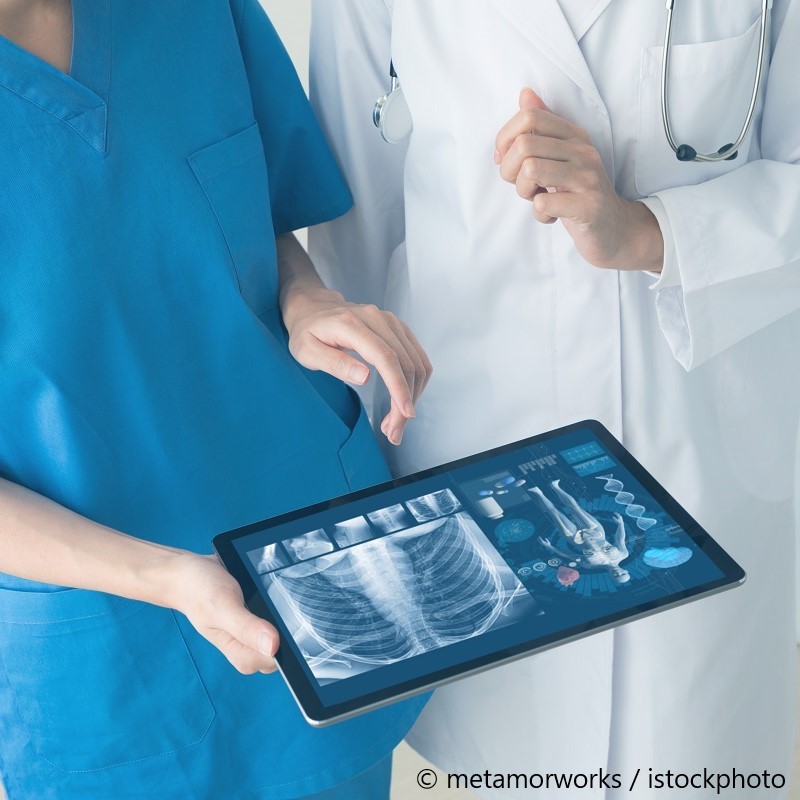 Zwei medizinische Fachkräfte betrachten ein Tablet. Auf dem Bildschirm sind Röntgenbilder sowie medizinische Grafiken zu sehen, die anatomische Informationen darstellen. Die Person in blauer Kleidung zeigt auf eine Stelle des Tablets, während die andere Person aufmerksam zuhört.