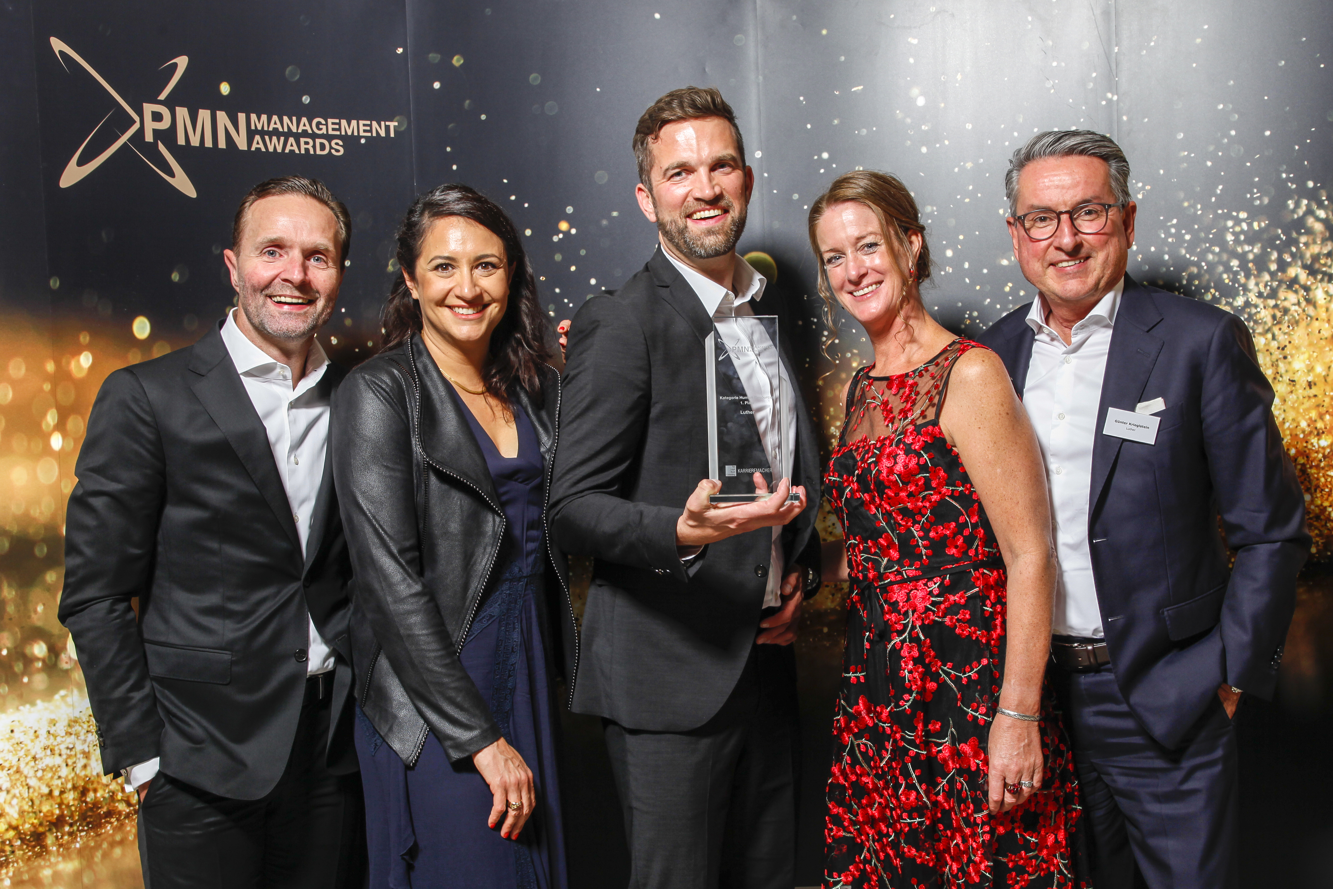 A group of five people stands together, smiling and posing for a photo. One person in the center holds an award. They are dressed in formal attire against a backdrop with a sparkling theme, suggesting a celebration or awards ceremony.