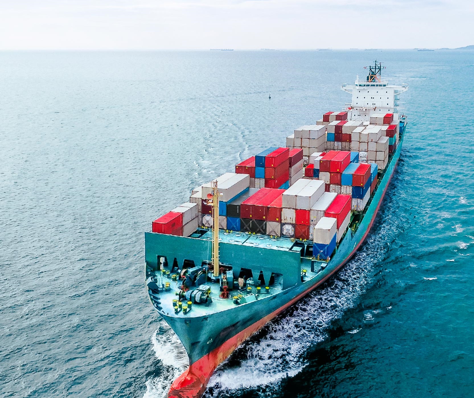 An aerial view of a large cargo ship navigating through open water, prominently loaded with colorful shipping containers. The ship's stern trails a wake as it moves, showcasing its journey across the sea.