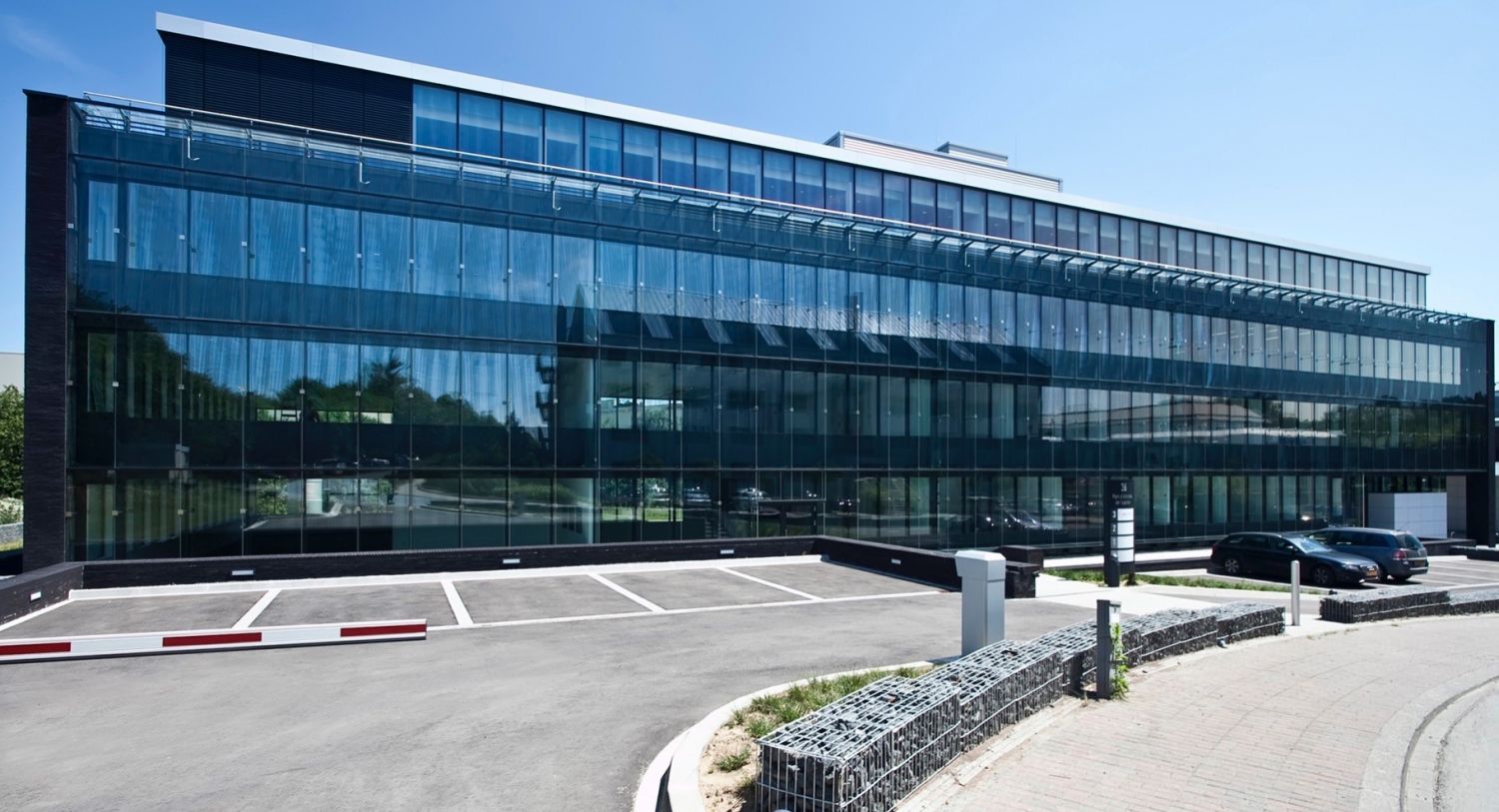 A modern, multi-story building with large glass windows reflects the blue sky. The structure features a sleek design and is set in a paved area with parking spaces visible in front. Greenery can be seen around the property, adding a touch of nature to the urban setting.
