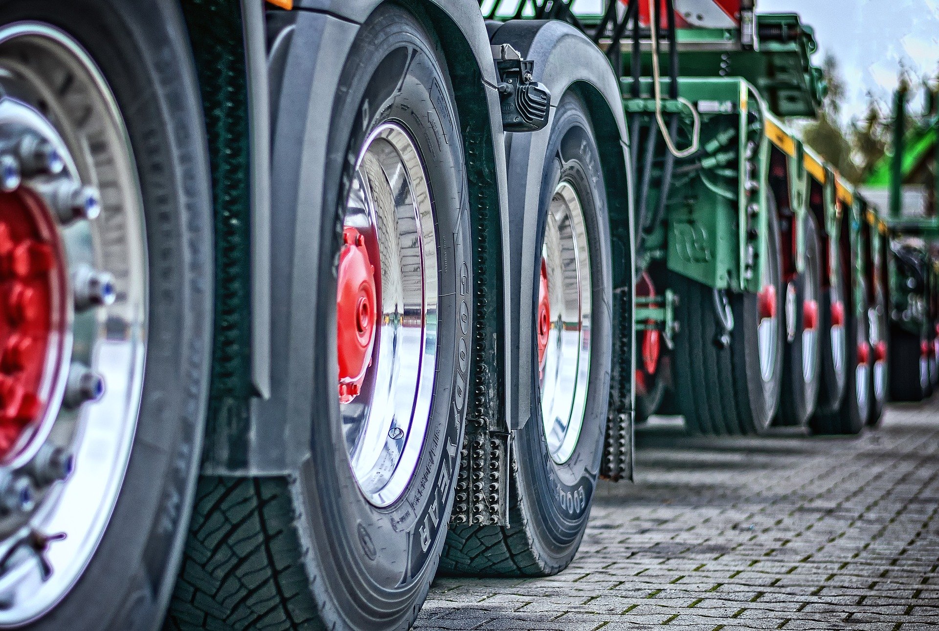 Eine Reihe von Lkw-Reifen, die im Fokus stehen. Die Felgen sind silber mit roten Mitten und die Reifen haben ein deutliches Profil. Der Untergrund besteht aus Pflastersteinen, die in einem gleichmäßigen Muster verlegt sind.