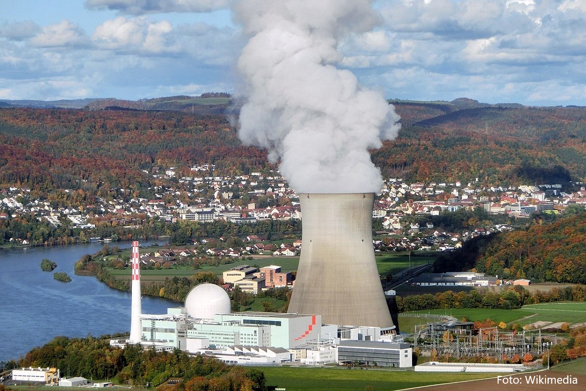Ein Kernkraftwerk mit einem großen Kühlturm, aus dem Dampf aufsteigt, ist im Vordergrund zu sehen. Im Hintergrund erstreckt sich eine Landschaft mit bunten Bäumen und einem Fluss, während sich Gebäude und Häuser am Ufer entlangreihen.